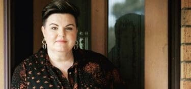 Photo of Kristy Forbes standing in a doorway wearing earrings and a patterned shirt.