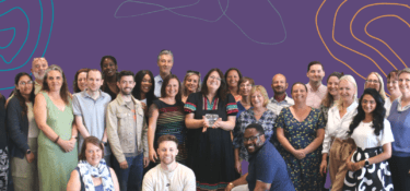 Photo of a diverse group of Genius Within staff all smiling at the camera. The background is dark purple with colourful wavy lines around the edges.
