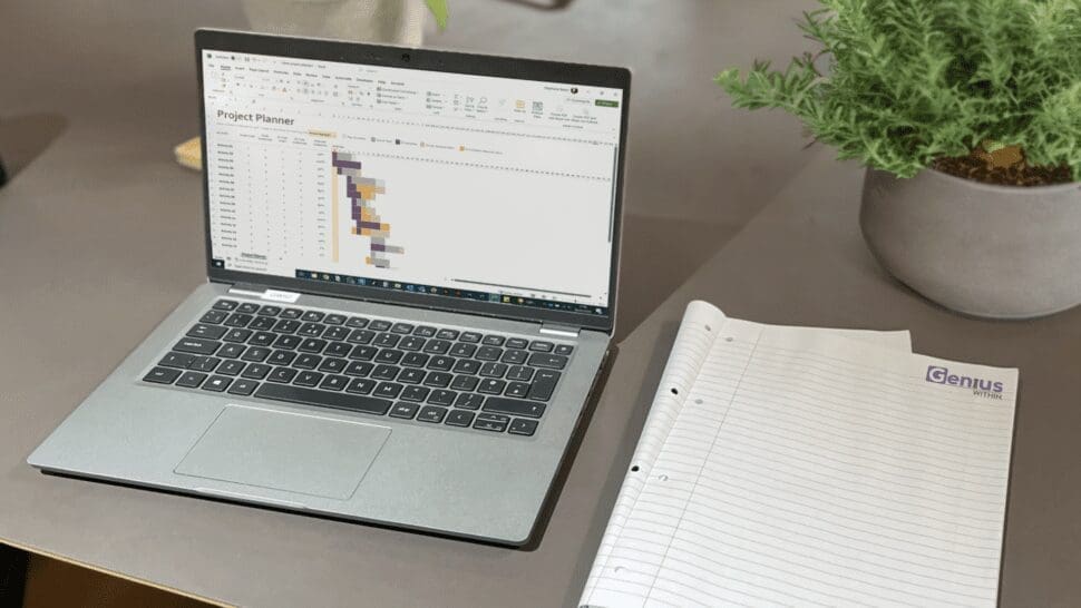 A laptop is open on a desk showing a project plan Gantt chart. There is an open note book next to he laptop with Genius Within's Logo in the top right corner.