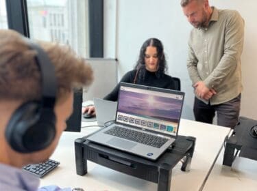 two men and a woman working at desks, one man is standing and helping the woman complete her neurodiversity awareness eLearning.