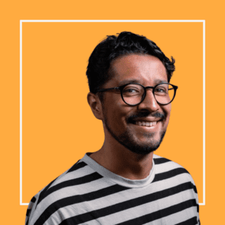 Zaq Mughal against a yellow background, wearing a stripey black and white top and glasses, smiling at the camera.